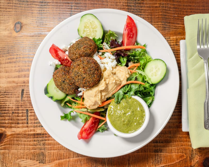 Falafel salad with hummus, feta, tomatoes, cucumbers, and green dressing on a white plate.