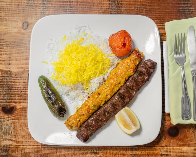 Two skewers of kebab, white and yellow rice, a grilled tomato, and a pepper on a white plate.