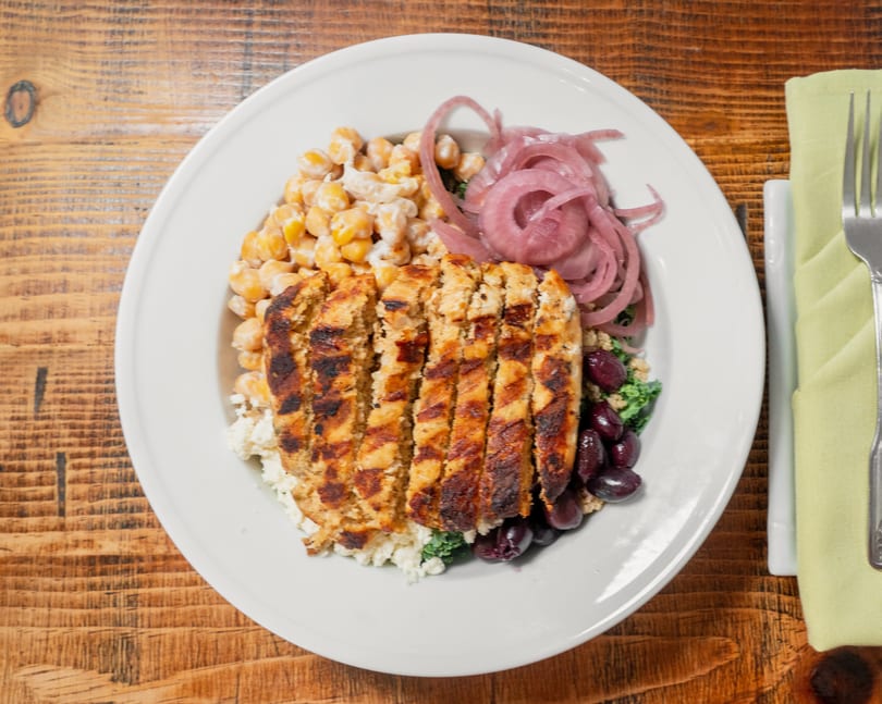 Grilled chicken, chickpeas, pickled red onions, and olives in a white bowl on a wooden table.