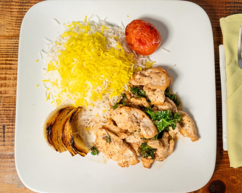 Chicken and kale with white and yellow rice, grilled onion, and a roasted tomato on a white plate.