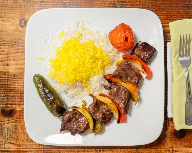 A plate of beef and vegetable skewers with white and yellow rice, a roasted tomato, and a pepper.
